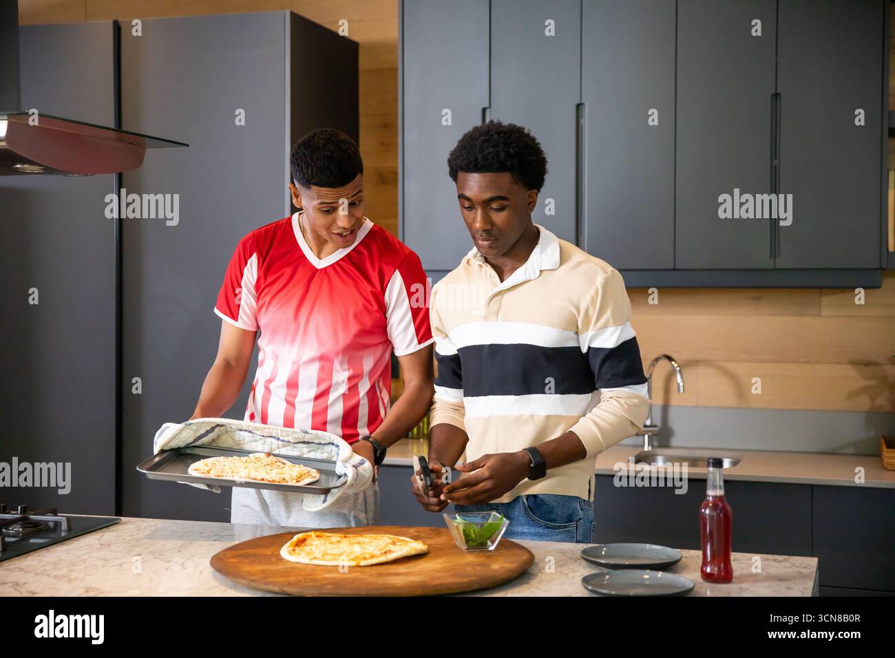 Diversi amici maschi preparano la pizza nella moderna cucina casalinga utilizzando vassoio, guanto da forno ed erbe fresche Foto Stock