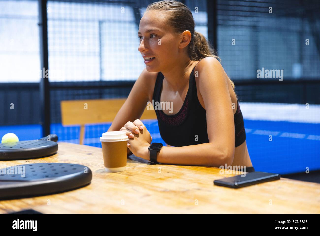 Donna che si prende una pausa dopo la partita di padel, seduta al tavolo accanto al campo di padel con racchette e tazza di caffè Foto Stock