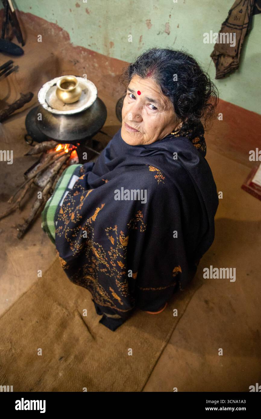 Un'anziana donna indiana siede accanto a una stufa tradizionale con una pentola di ottone in una casa rustica. La calda scena mette in risalto la vita quotidiana, la famiglia, la cura e l'endurina Foto Stock