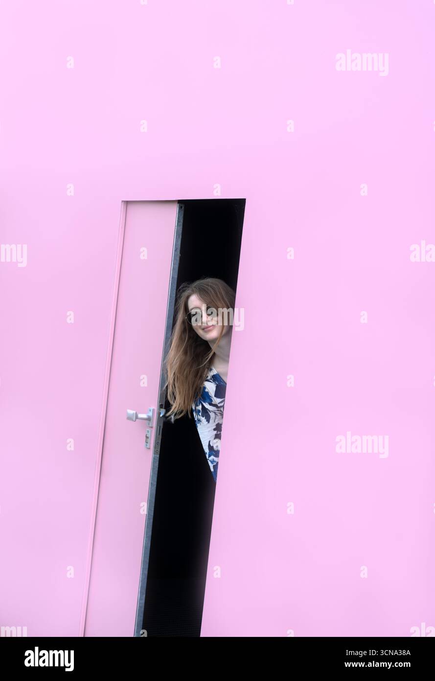 Una donna sorridente con occhiali da sole sbuca fuori da una porta rosa parzialmente aperta, inondata di luce soffusa. Foto Stock