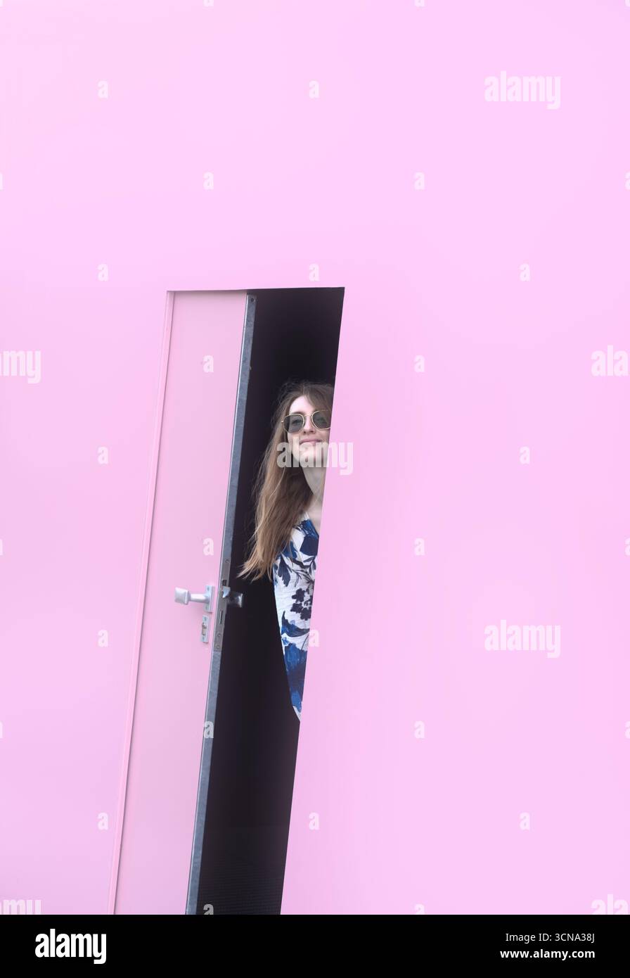 Una donna sorridente con occhiali da sole sbuca fuori da una porta rosa parzialmente aperta, inondata di luce soffusa. Foto Stock