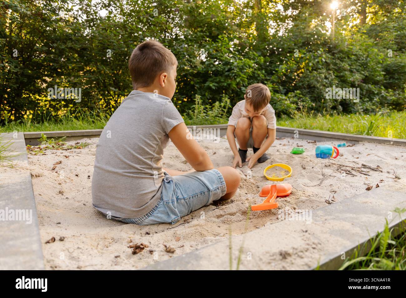 Due bambini giocano in un'area sabbiosa, costruendo e scavando in modo creativo mentre si godono il sole. La vegetazione lussureggiante che circonda contribuisce al divertimento all'aria aperta, cr Foto Stock