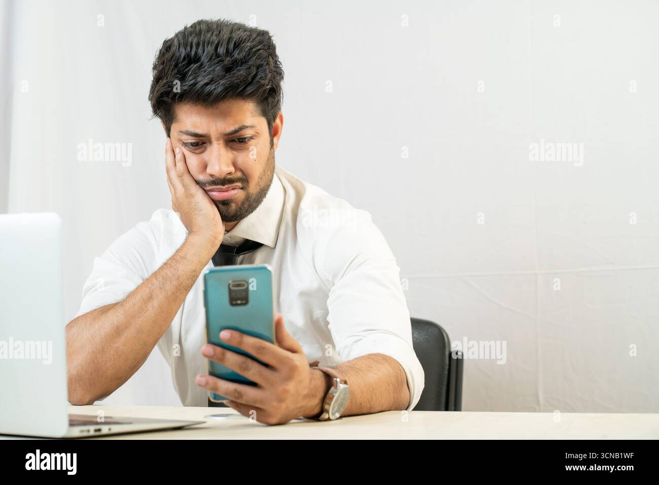 Un giovane uomo a una scrivania con un portatile, che sembra annoiato e triste al telefono. Foto Stock