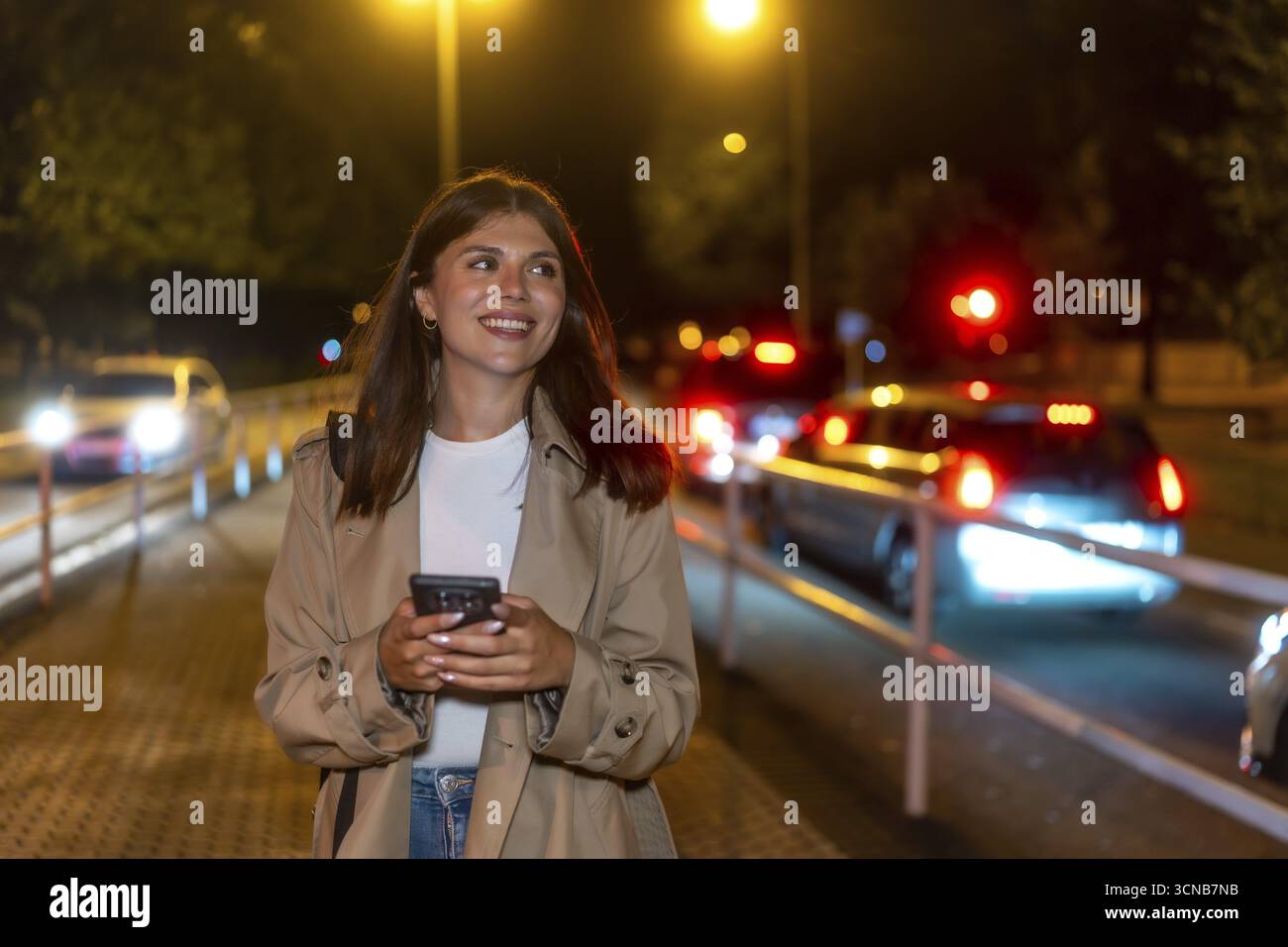 Giovane donna con un trench sorridente, con un telefono in mano e con lo sguardo lontano mentre cammina su una strada della città di notte con luci urbane e traffico sfocato Foto Stock