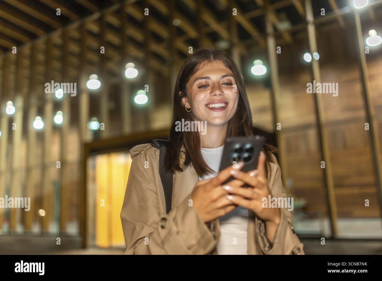 Giovane donna in un trench coat sorridente, che controlla il suo smartphone e vive momenti felici mentre naviga nella vita moderna della città, illuminata dall'atmosfera Foto Stock