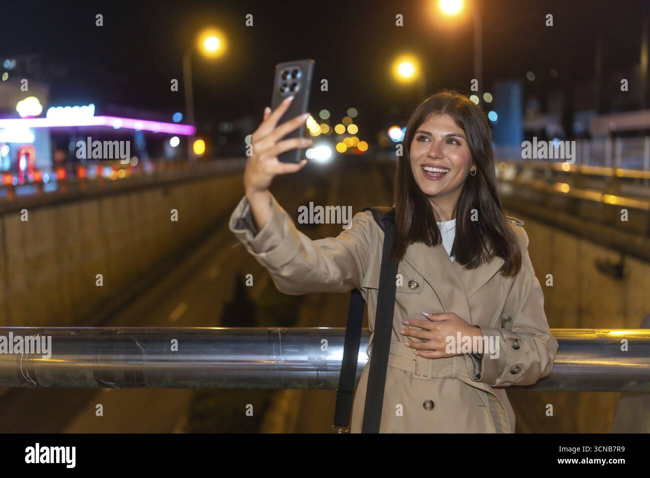 Giovane donna sorridente e scattata un selfie con uno smartphone di notte, catturando momenti di connessione e vita urbana su un ponte con le luci della città nel Foto Stock