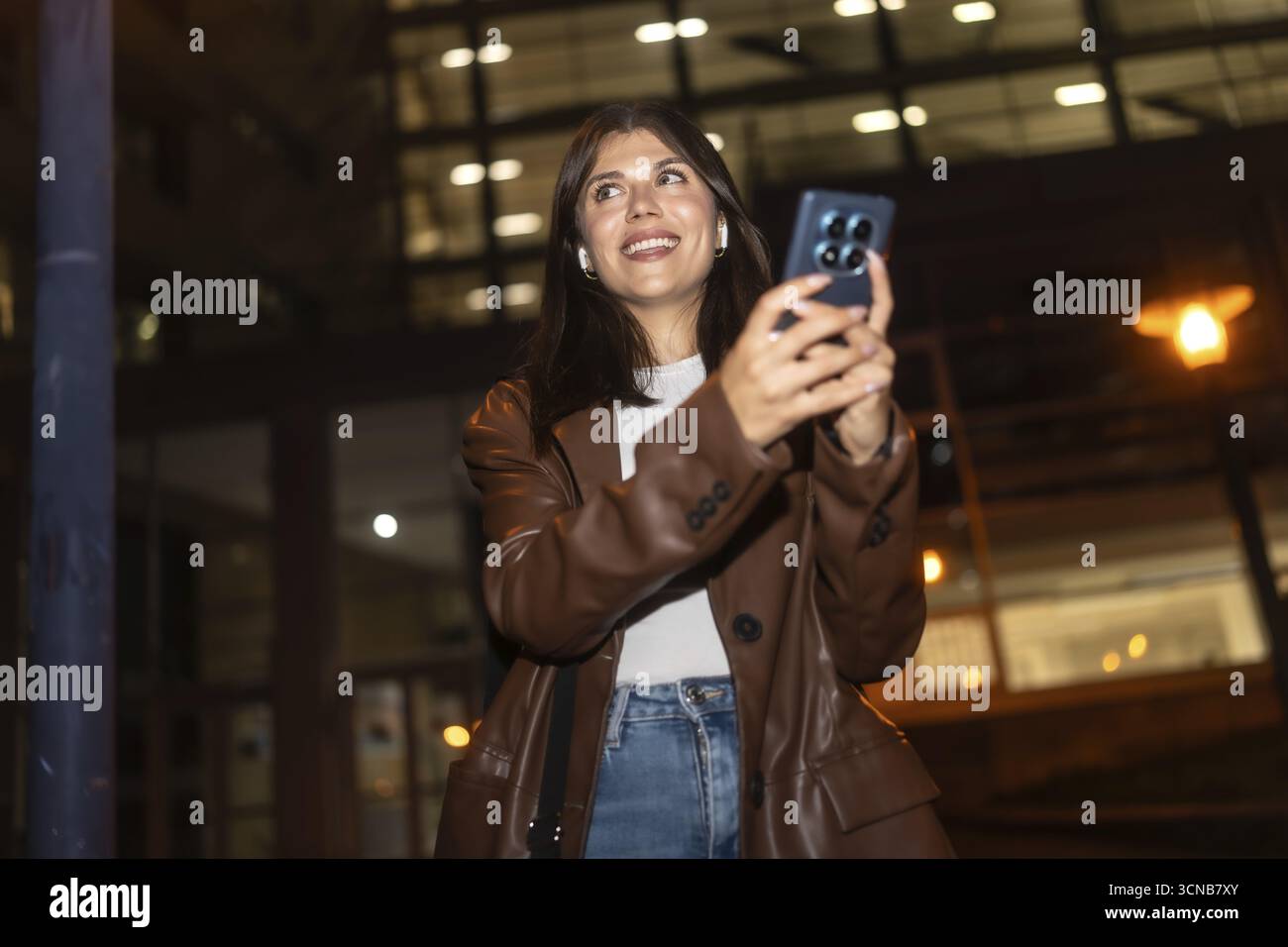 La giovane donna sorride e guarda in alto mentre utilizza lo smartphone e gli auricolari wireless di notte, cattura i momenti urbani o comunica con il ven Foto Stock