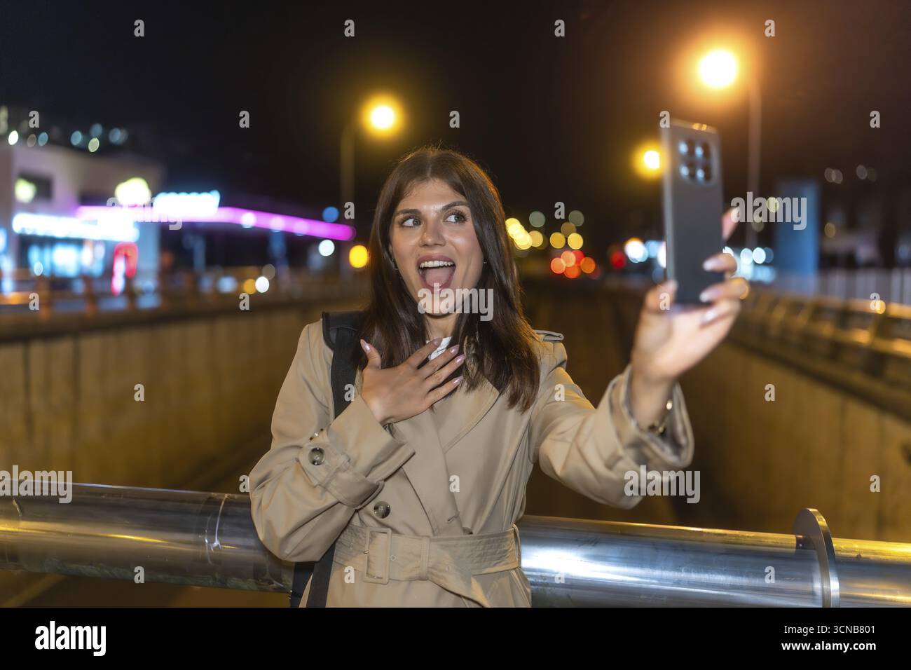 Giovane donna in trench coat sorpresa e gioiosa scattando un selfie notturno in una vivace strada cittadina, con un telefono che si illumina contro luci sfocate per il vlogging Foto Stock