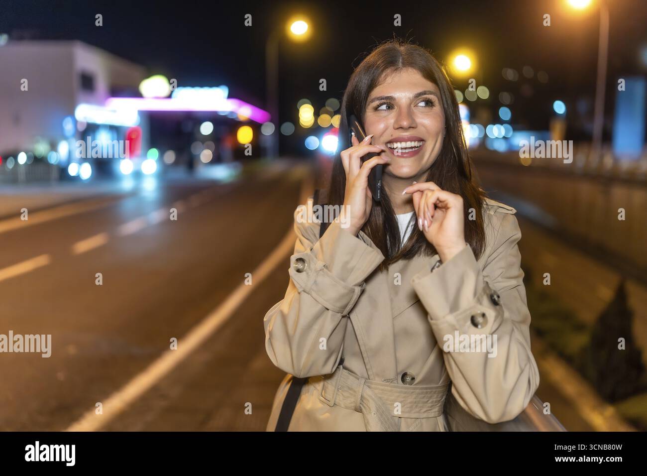 Una giovane donna in un trench coat sorride e ride mentre parla con il suo smartphone durante una passeggiata notturna illuminata della città, godendosi un'atmosfera rilassata e gioiosa Foto Stock