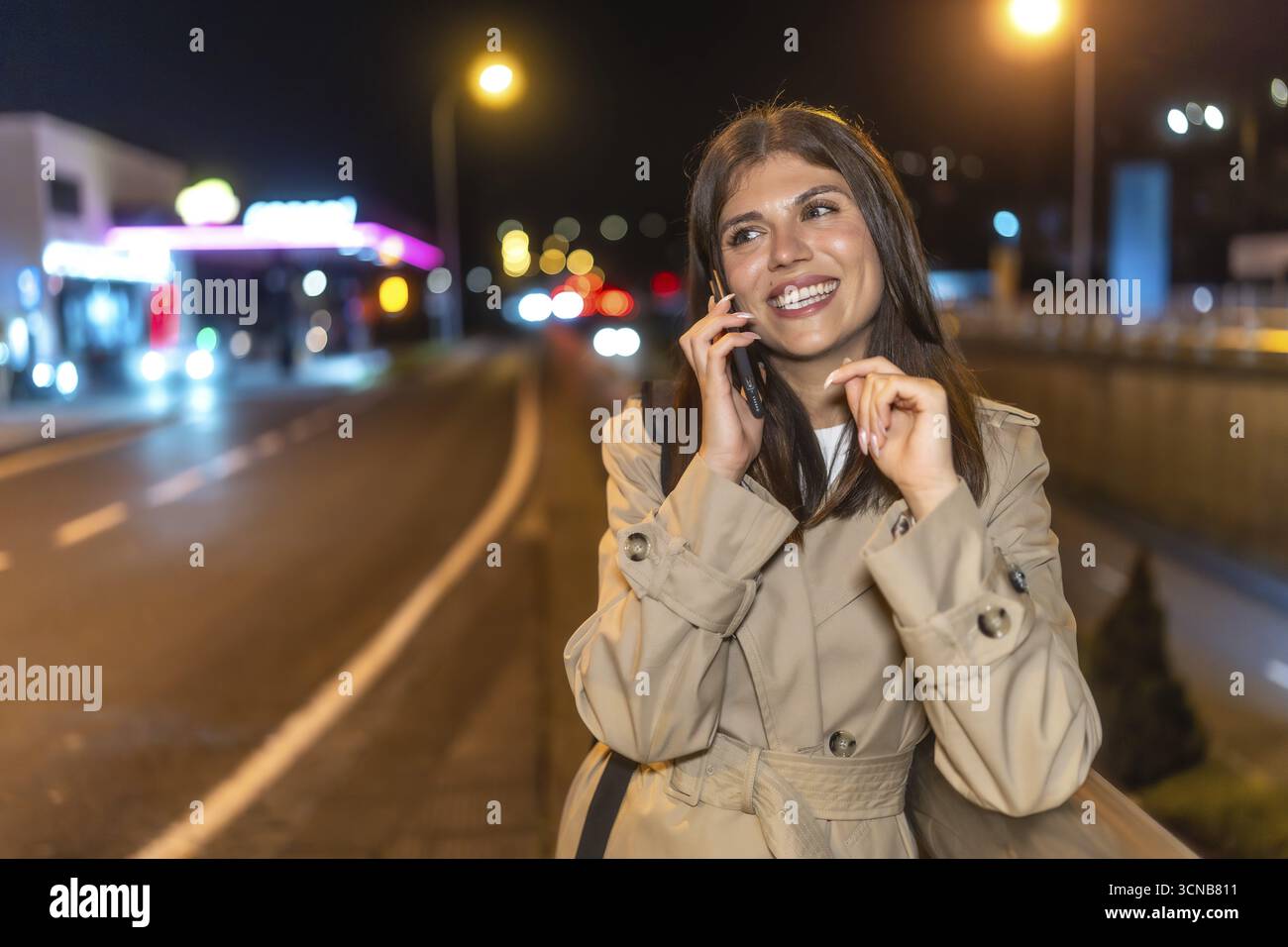 Giovane donna sorridente in un trench mentre parla al telefono cellulare in una vivace strada della città di notte, immersa nelle calde luci urbane e nel morbido bokeh b Foto Stock