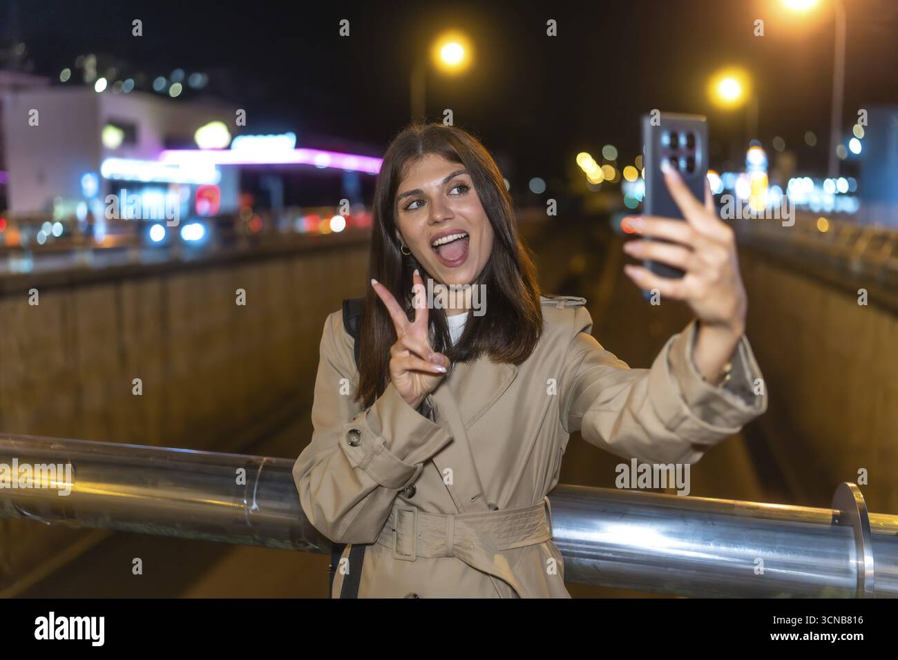 Giovane donna che indossa un trench in piedi su un ponte, regge uno smartphone e scatta un autoritratto, mostrando il segno di pace con un espresso felice Foto Stock