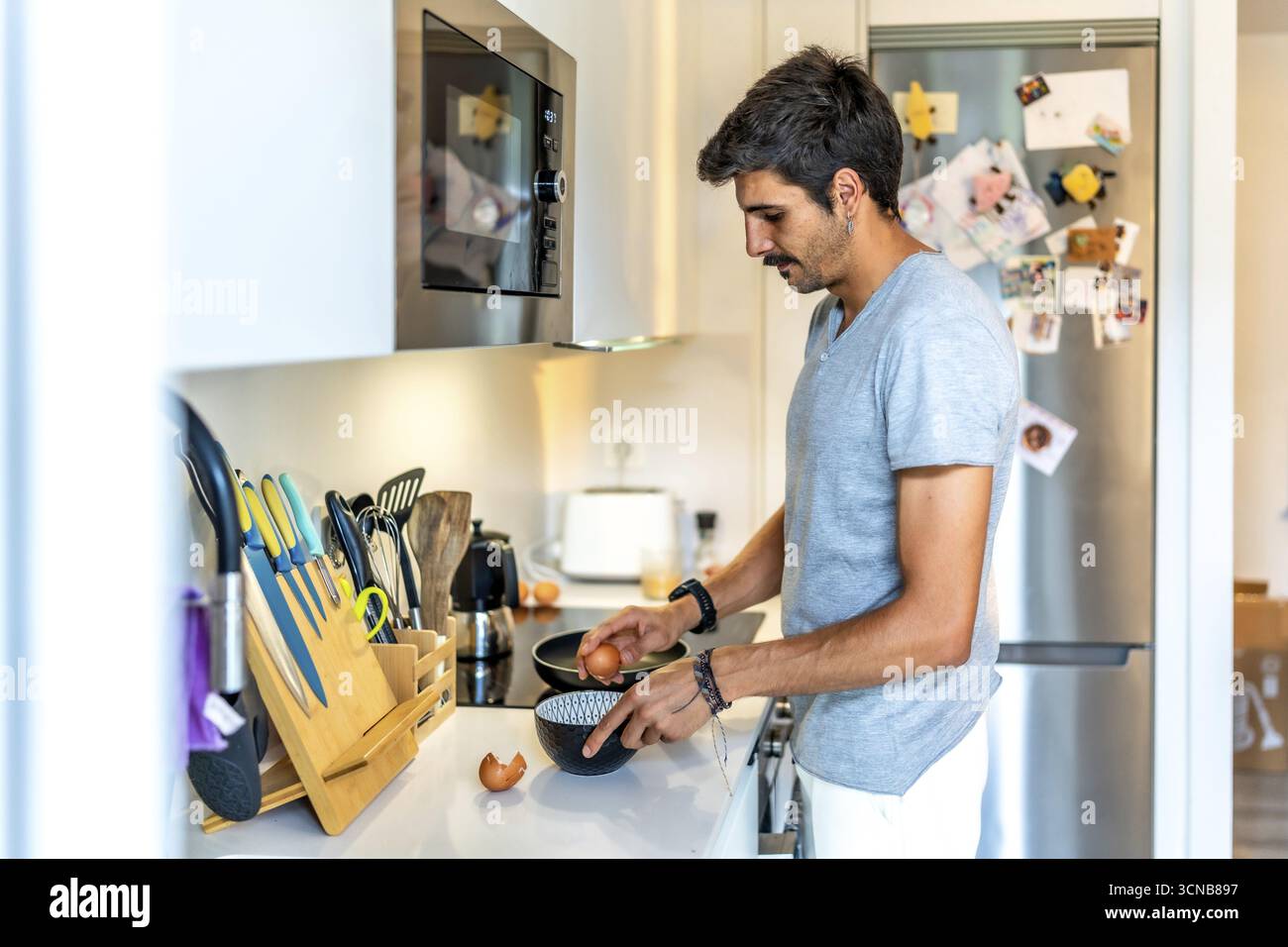 Il giovane spaccando le uova in una ciotola, preparandosi a cucinare la colazione o un pasto nella sua cucina moderna e ben attrezzata Foto Stock