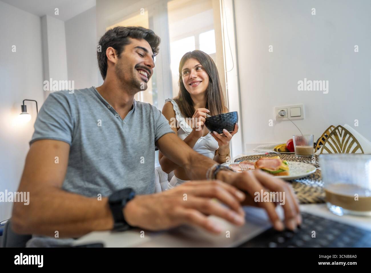 Una giovane coppia che fa colazione in una cucina luminosa, lavora da casa su un laptop e condivide sorrisi e conversazioni insieme Foto Stock