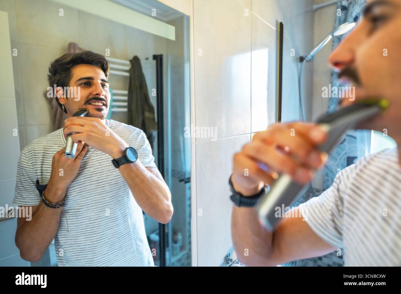 Giovane uomo che rifinisce la barba con un rasoio elettrico, guardandosi nello specchio del bagno durante la sua routine mattutina Foto Stock