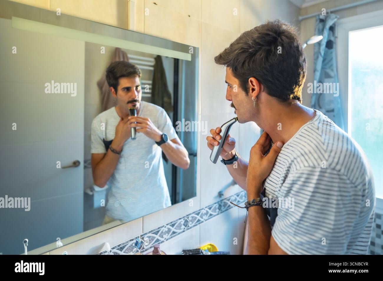 Uomo caucasico che rifilava i baffi con un rasoio elettrico mentre guardava nello specchio del bagno durante la sua routine mattutina Foto Stock