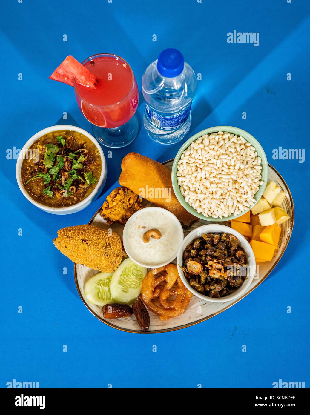 Uno scatto dall'alto di un tradizionale piatto di Iftar dell'Asia meridionale su sfondo blu. Il piatto include piatti dolci e salati, una bevanda e una bottiglia Foto Stock