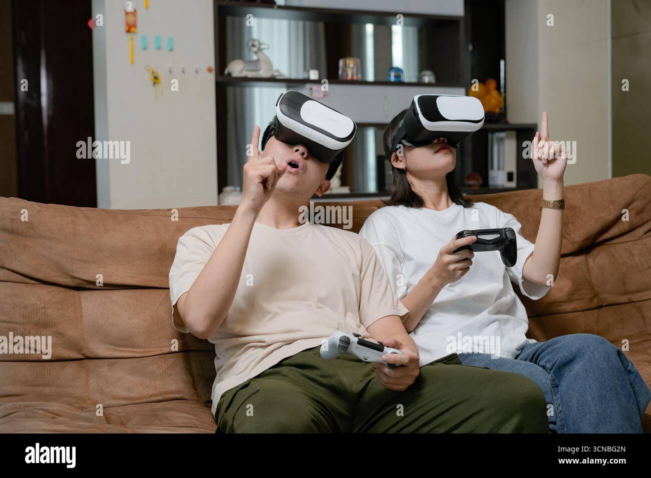 Due amici si rilassano sul divano di casa, indossando occhiali VR e utilizzando i controller per esplorare mondi virtuali immersivi Foto Stock
