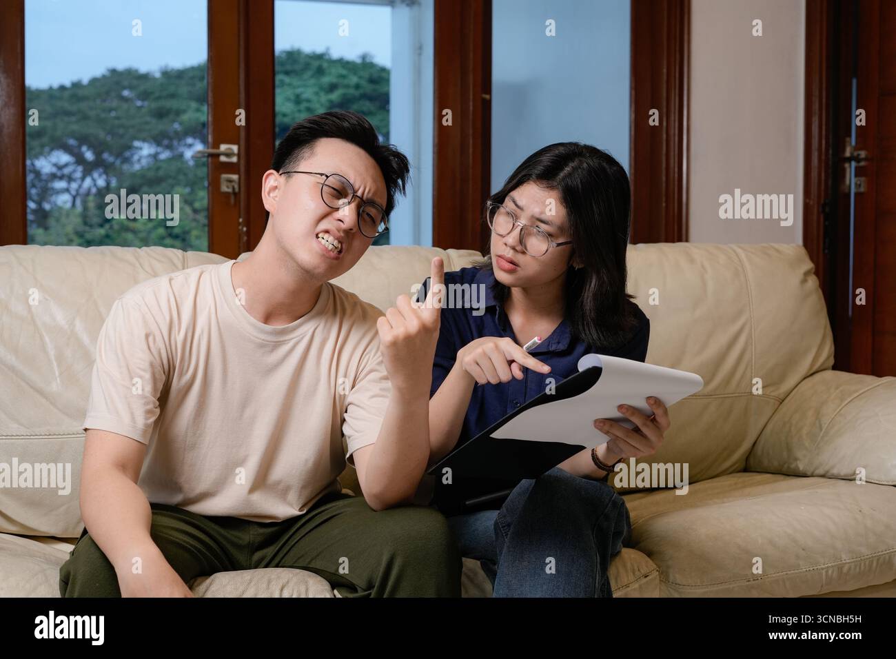 Un uomo ansioso siede su un divano beige mentre una donna nelle vicinanze esamina documenti e note, offre una guida e un supporto ponderati in un ambiente tranquillo Foto Stock