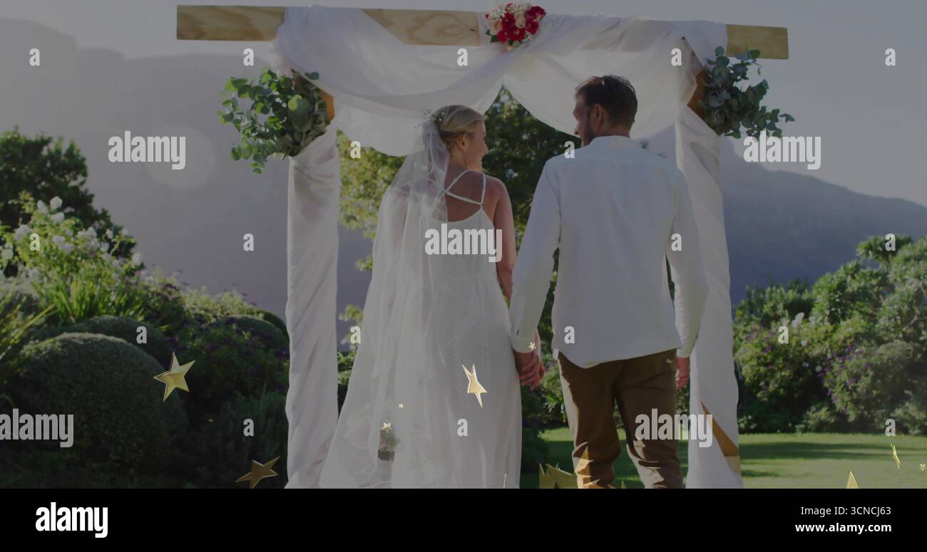 Sposa e sposo in piedi che tengono per mano sotto l'arco nuziale in giardino, con tendaggi, fiori, velo Foto Stock