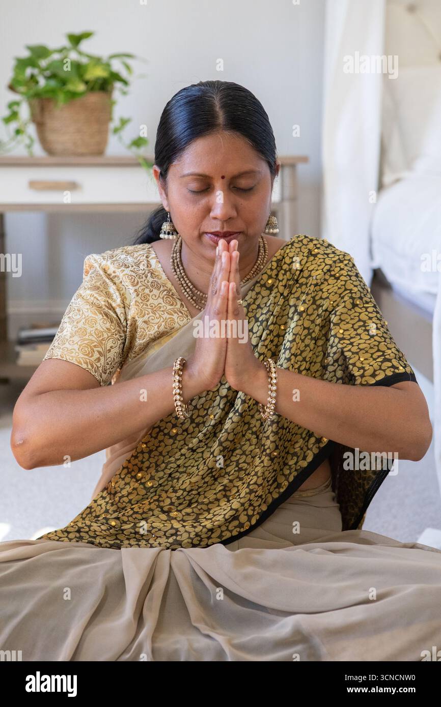 Donna indiana anziana che indossa sari dorati e neri che prega in camera da letto con piante in vaso Foto Stock