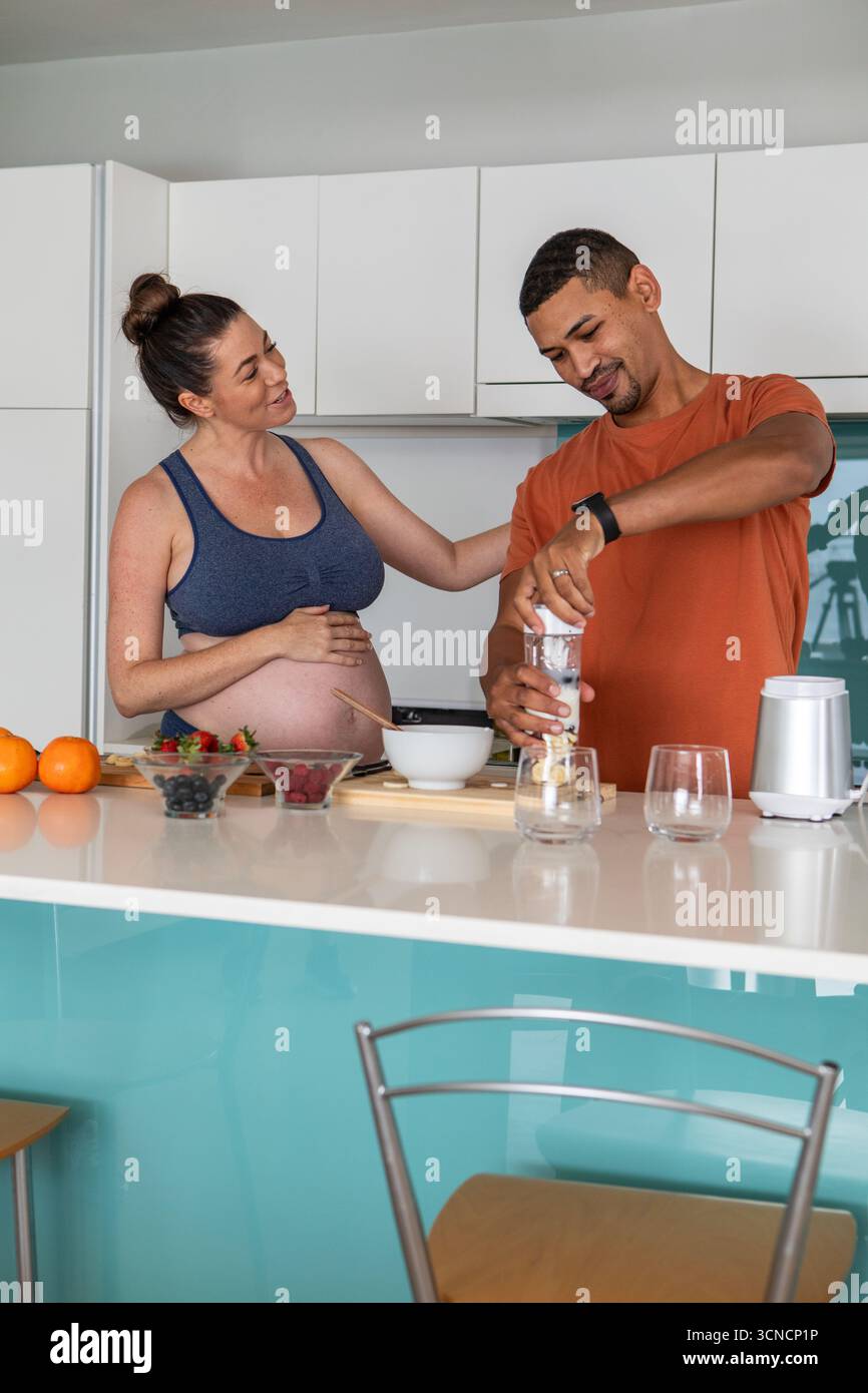 Coppia che indossa un abbigliamento sportivo e una maglietta dietro il bancone della cucina che mescola frutti di bosco e arance nel frullatore Foto Stock