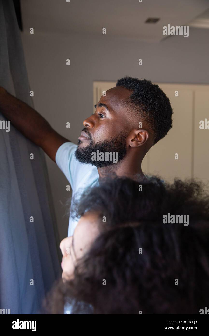 Coppia afroamericana in piedi accanto alla finestra tirando indietro le tende a casa, bocchetta di ventilazione sul soffitto Foto Stock