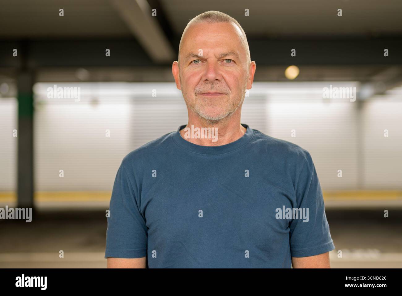 Uomo maturo con acconciatura corta e t-shirt blu, che guarda con sicurezza la fotocamera in un ambiente urbano con illuminazione soffusa. Foto Stock