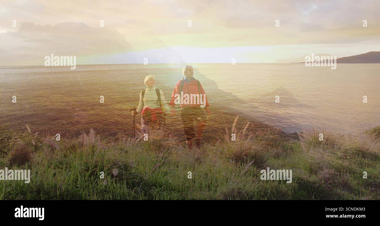 Una coppia di escursionisti che tengono in mano pali e zaini sulla scogliera erbosa della costa al tramonto, con un caldo bagliore Foto Stock