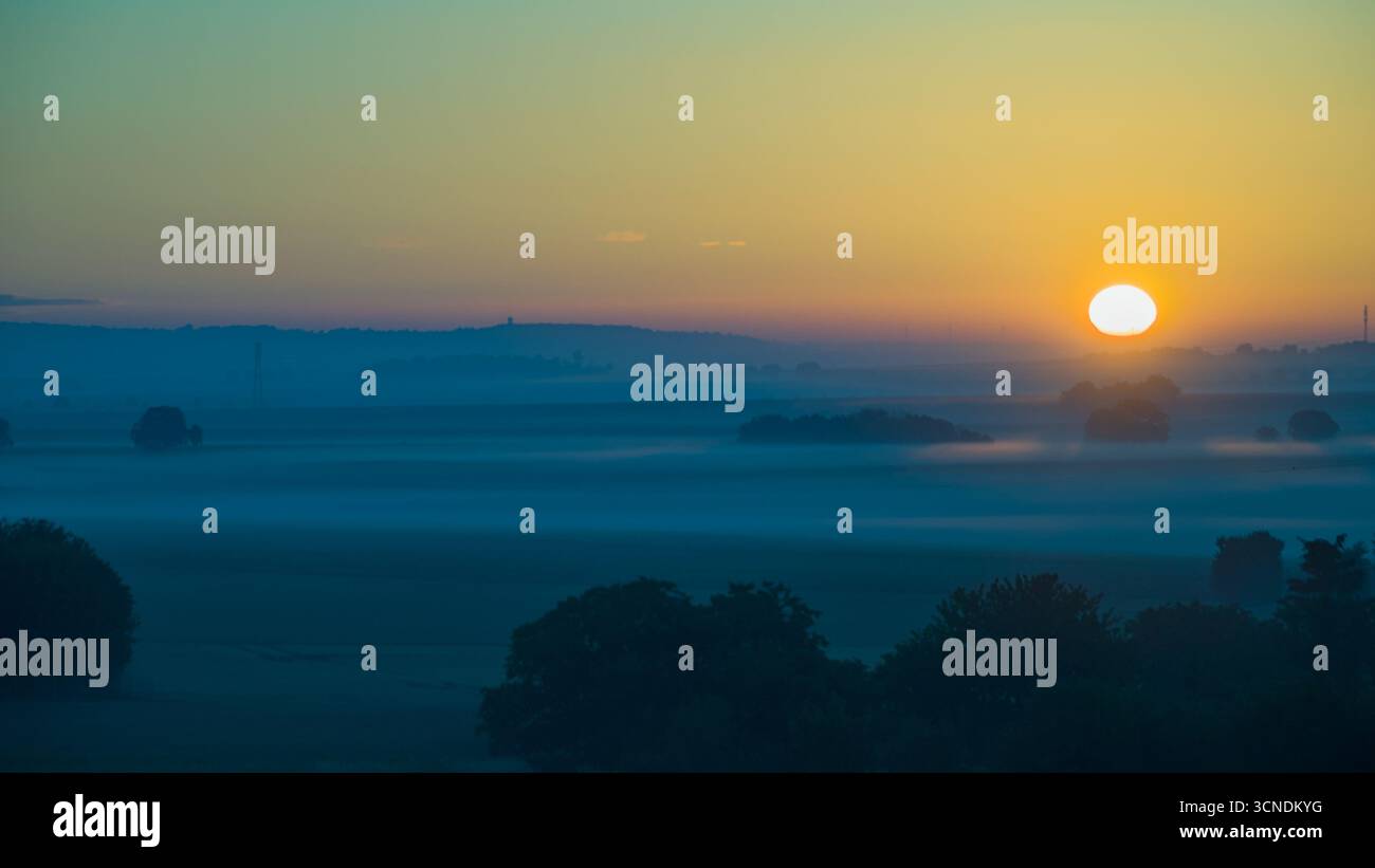 Alba sulla campagna francese con campi ondulati, alberi sparsi e nebbia mattutina, immersa nella luce del sole mattutino Foto Stock