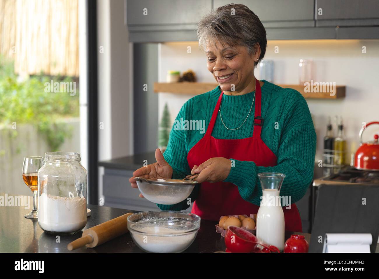 Donna afroamericana anziana che indossa grembiule rosso setacciando la farina attraverso il setaccio di metallo a Kitchen Island Foto Stock