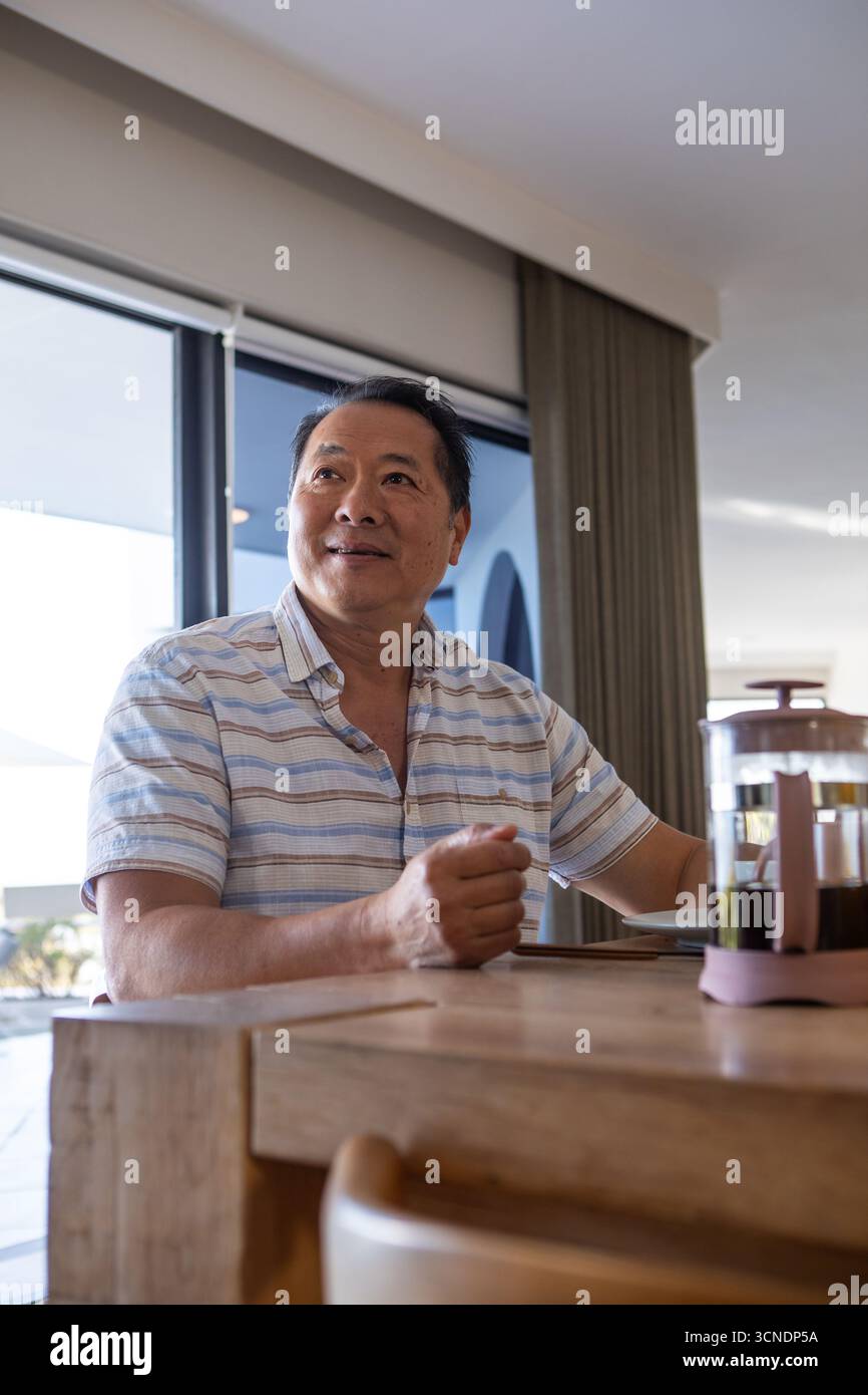 Uomo asiatico/cinese seduto al tavolo da pranzo nella zona pranzo, guardando a sinistra accanto alla stampa francese Foto Stock