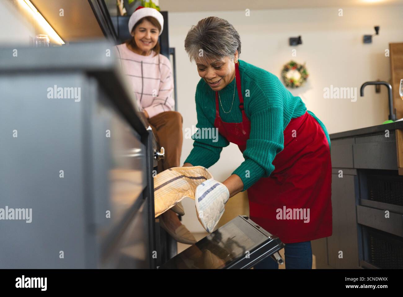 Diverse amiche che cucinano a casa, donna anziana che rimuove il piatto dal forno con guanto dalla corona Foto Stock