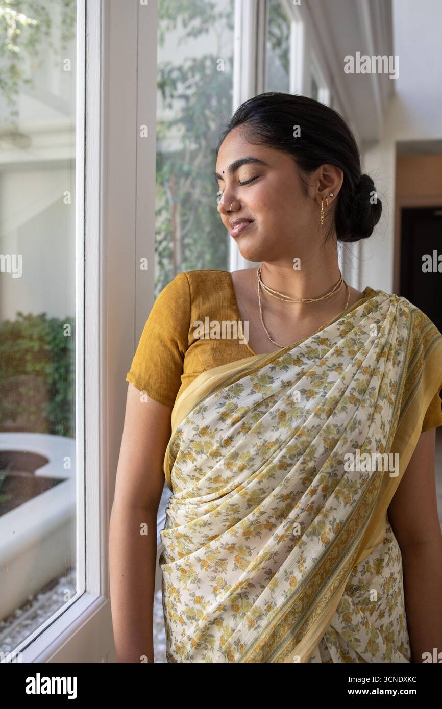 Donna indiana che indossa una camicetta di senape sari e gioielli in oro mentre è in piedi davanti a una finestra a casa Foto Stock