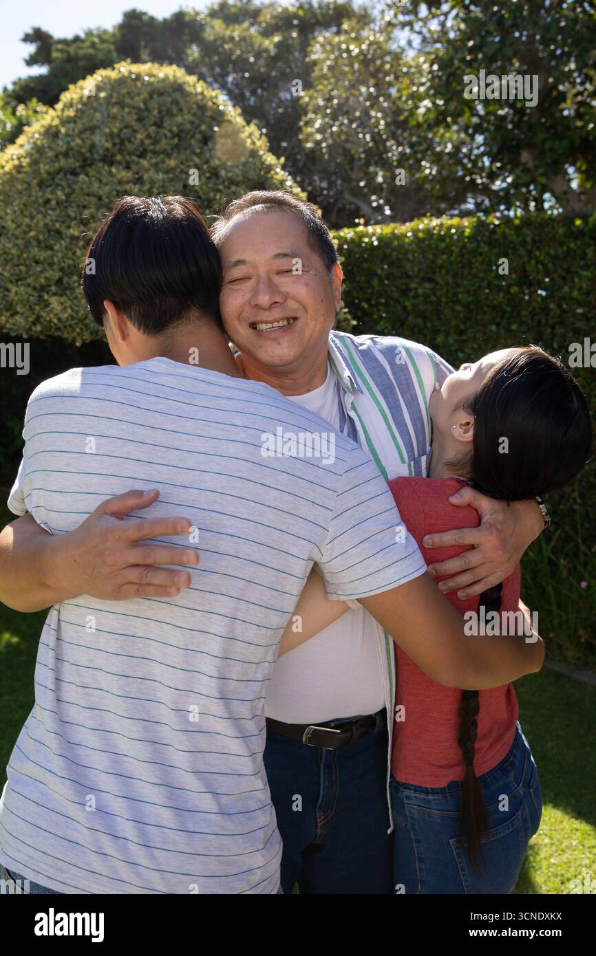 Famiglia asiatica che abbraccia sotto un arco di siepe scolpito in un giardino sul retro illuminato dal sole e indossa camicie a righe Foto Stock