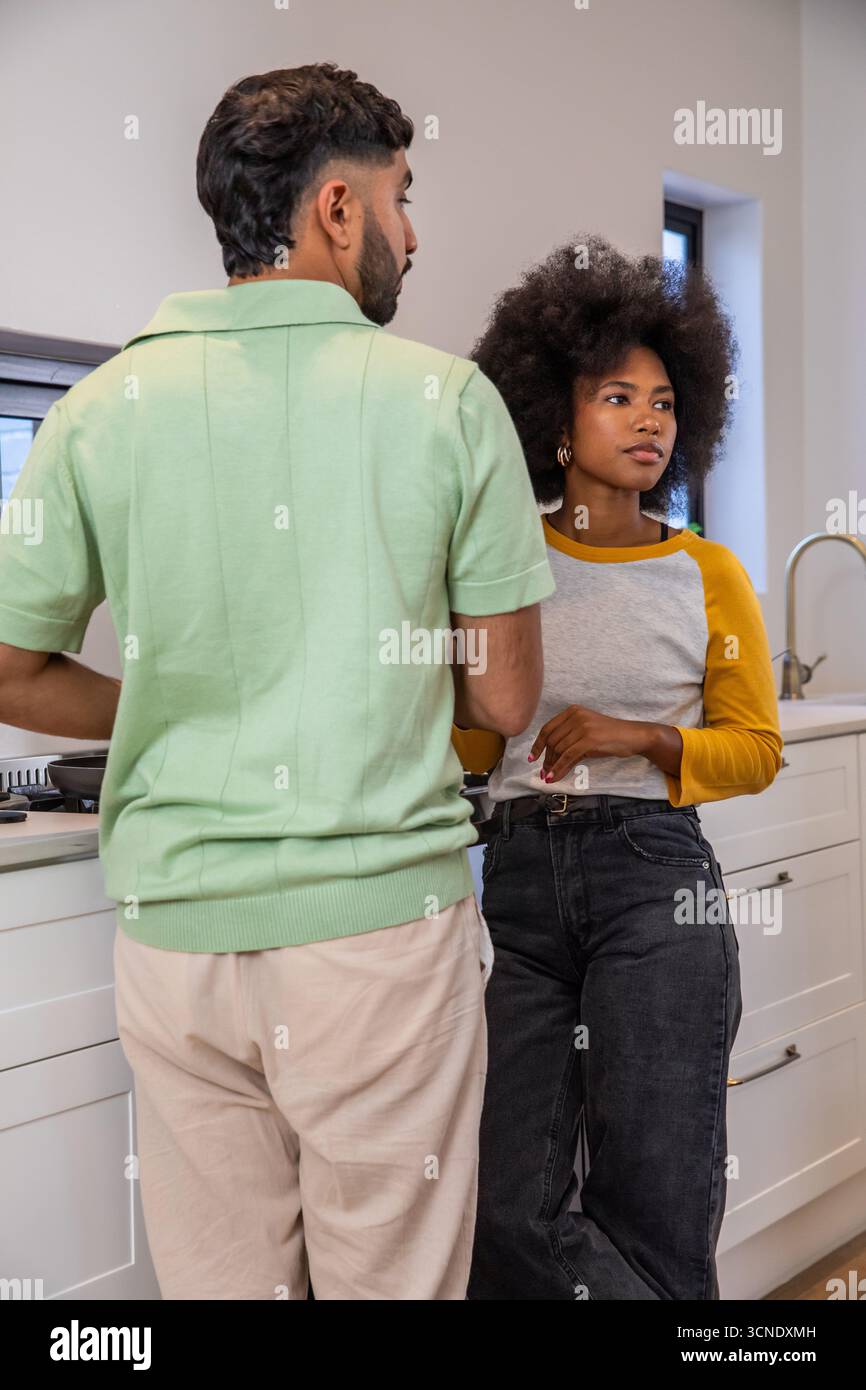 Coppia variegata in piedi sul piano cottura e appoggiata al bancone con utensili per preparare il pasto Foto Stock