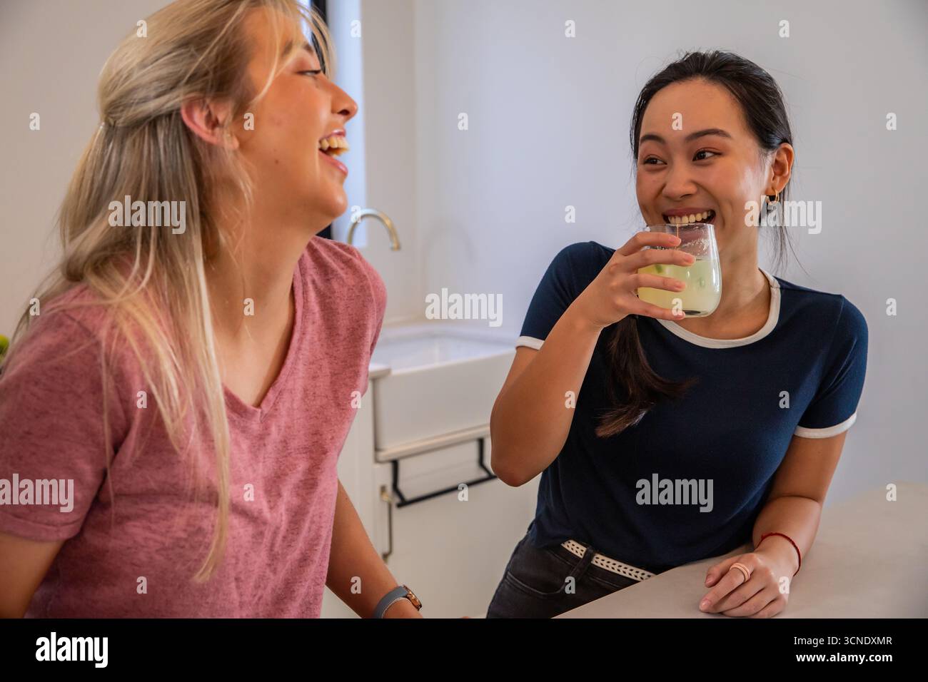 Diverse amiche che ridono e sorseggiano succo verde sul piano della cucina con rubinetto Foto Stock
