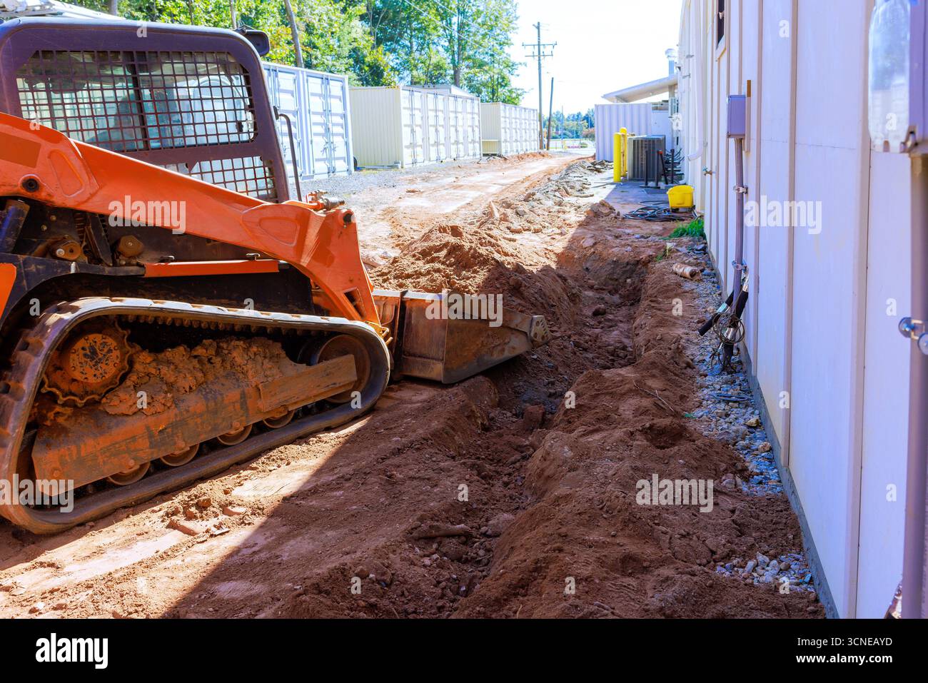 L'escavatore da cantiere scava fossati accanto a edifici in fase di lavorazione nell'area suburbana. Foto Stock