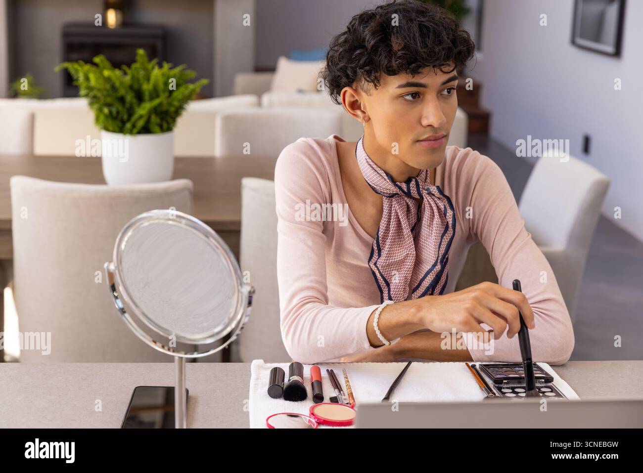 Uomo in cima rosa seduto a un tavolo di casa applicando trucco con pennelli, tavolozza, specchio Foto Stock