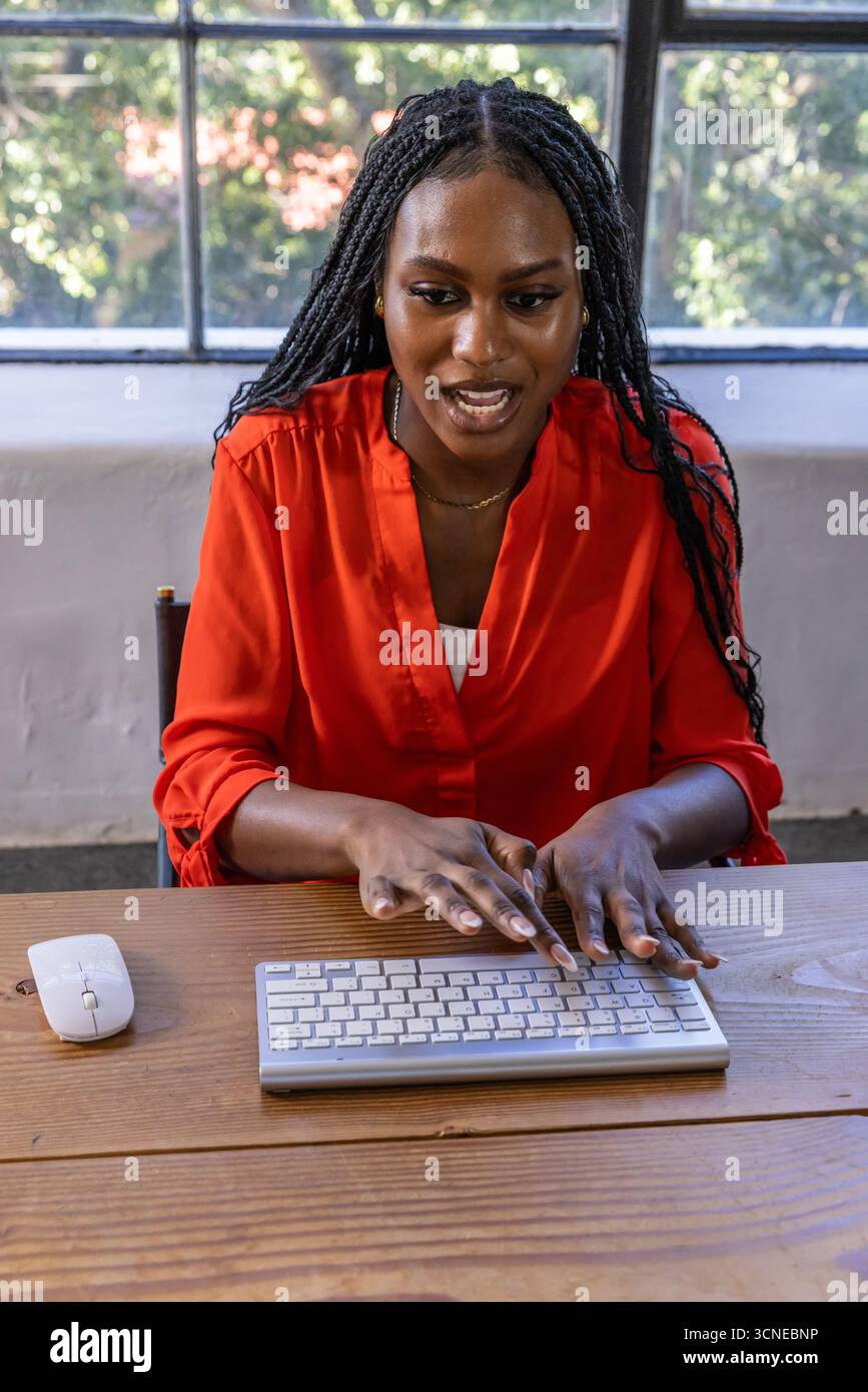 Donna afroamericana che digita con tastiera wireless, mouse alla scrivania dell'ufficio per finestra Foto Stock