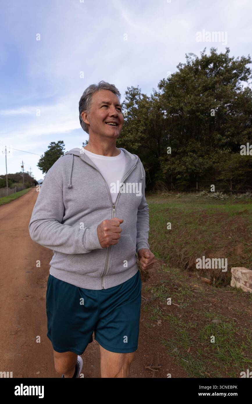 Uomo che fa jogging su strada sterrata indossando felpa con cappuccio, camicia girocollo, pantaloncini sportivi e scarpe da running Foto Stock