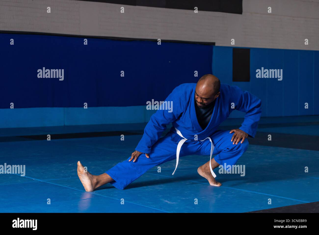 Uomo afro-americano in gi blu, cintura bianca che esegue affondamento laterale sui tappetini prima dei pannelli imbottiti Foto Stock
