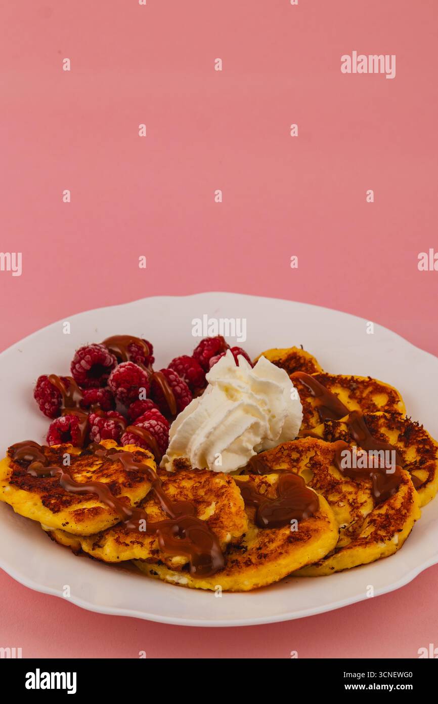 I pancake appena cucinati sono serviti con panna e salsa dolce, insieme a succosi lamponi. Il vivace sfondo rosa rende il piatto accattivante Foto Stock