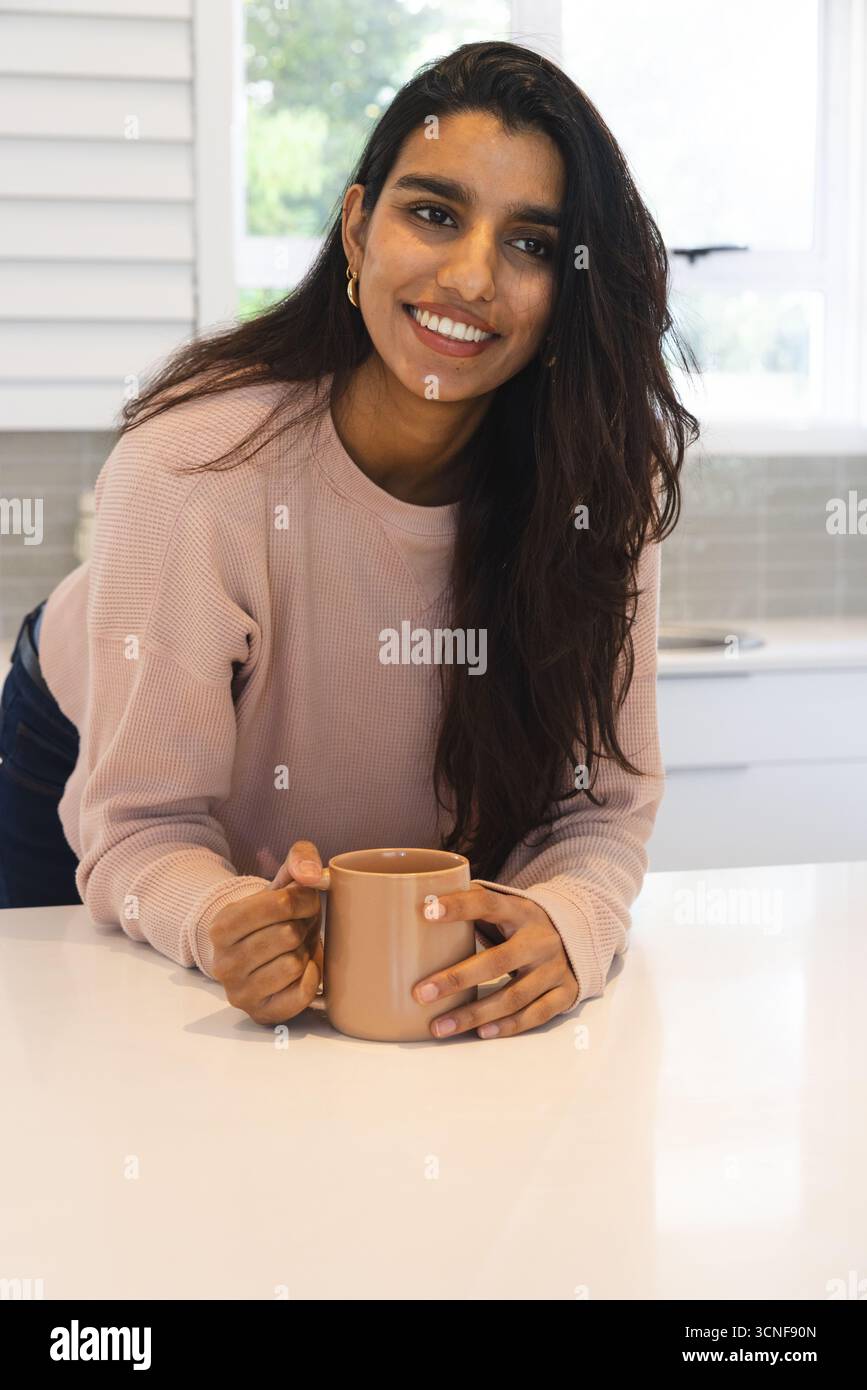 Donna dell'Asia meridionale appoggiata sul piano di lavoro in cucina mentre tiene in mano una tazza di ceramica abbronzata Foto Stock