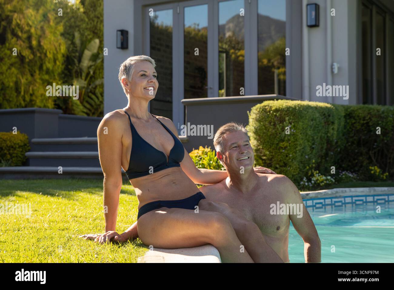 Coppia senior che si rilassa a bordo piscina nel cortile posteriore indossando bikini neri e costume da bagno accanto alla siepe Foto Stock