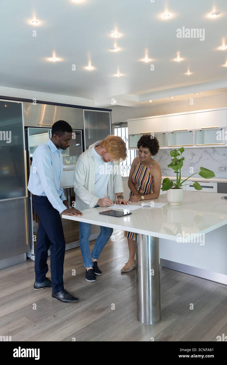Agente immobiliare diversificato, coppia che esamina i documenti di acquisto utilizzando un tablet a Kitchen Island Foto Stock