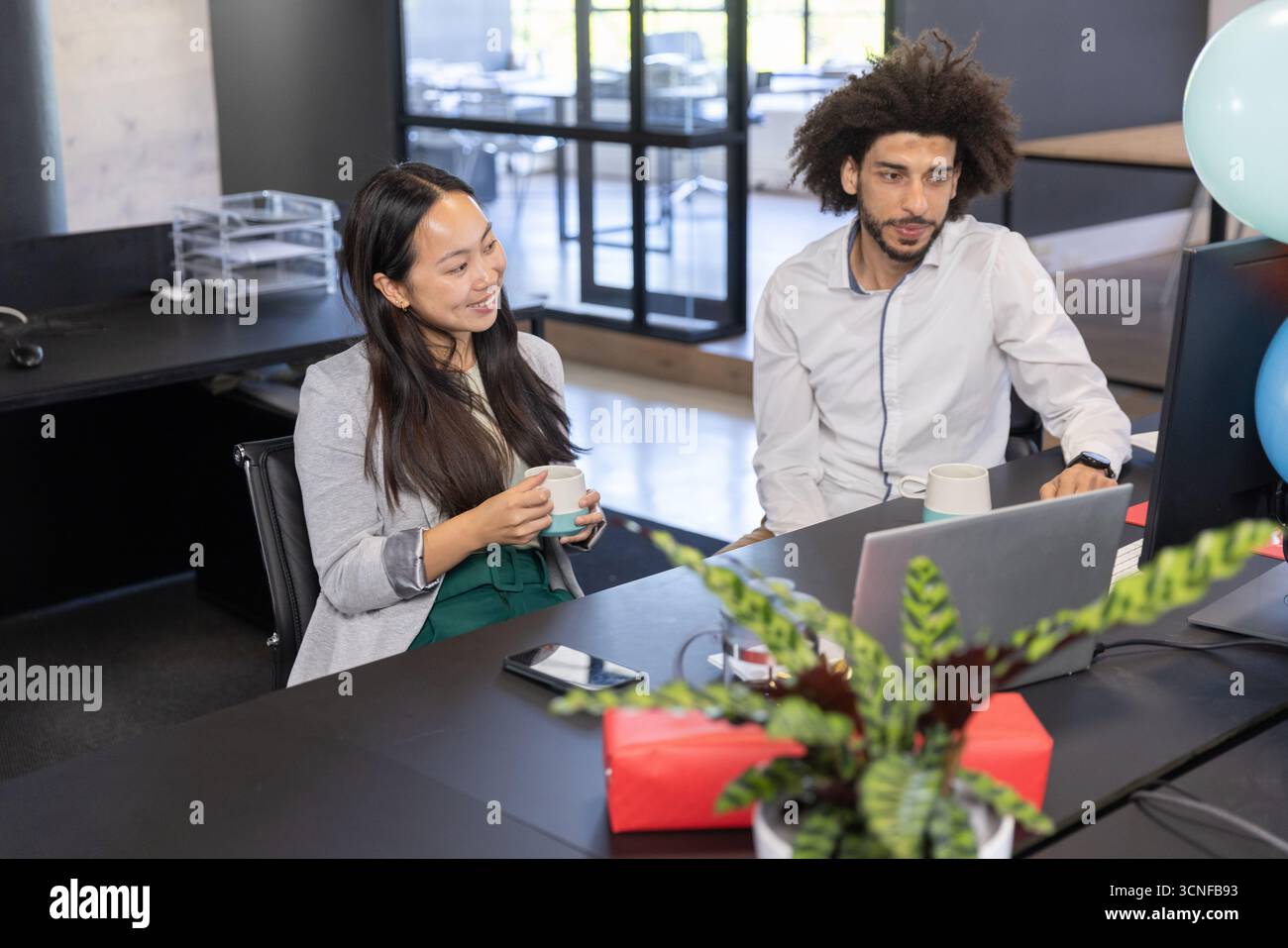 Colleghi diversi che collaborano alla scrivania nera con laptop, monitor e scatola regalo in ufficio Foto Stock