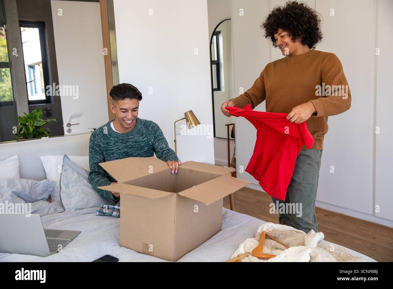 Diversi amici maschi che disimballano la scatola di cartone in camera da letto tenendo la camicia rossa accanto al computer portatile Foto Stock