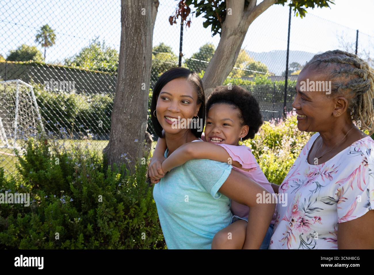 Famiglia variegata con nonna che guarda la madre che fa il piggybacking figlia vicino alla recinzione della catena nel parco Foto Stock