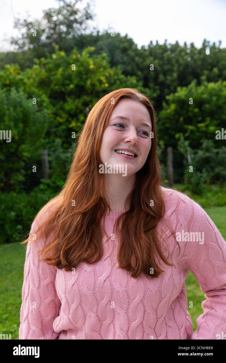 Donna sorridente dai capelli rossi in piedi nel giardino erboso sul retro con un maglione rosa Foto Stock