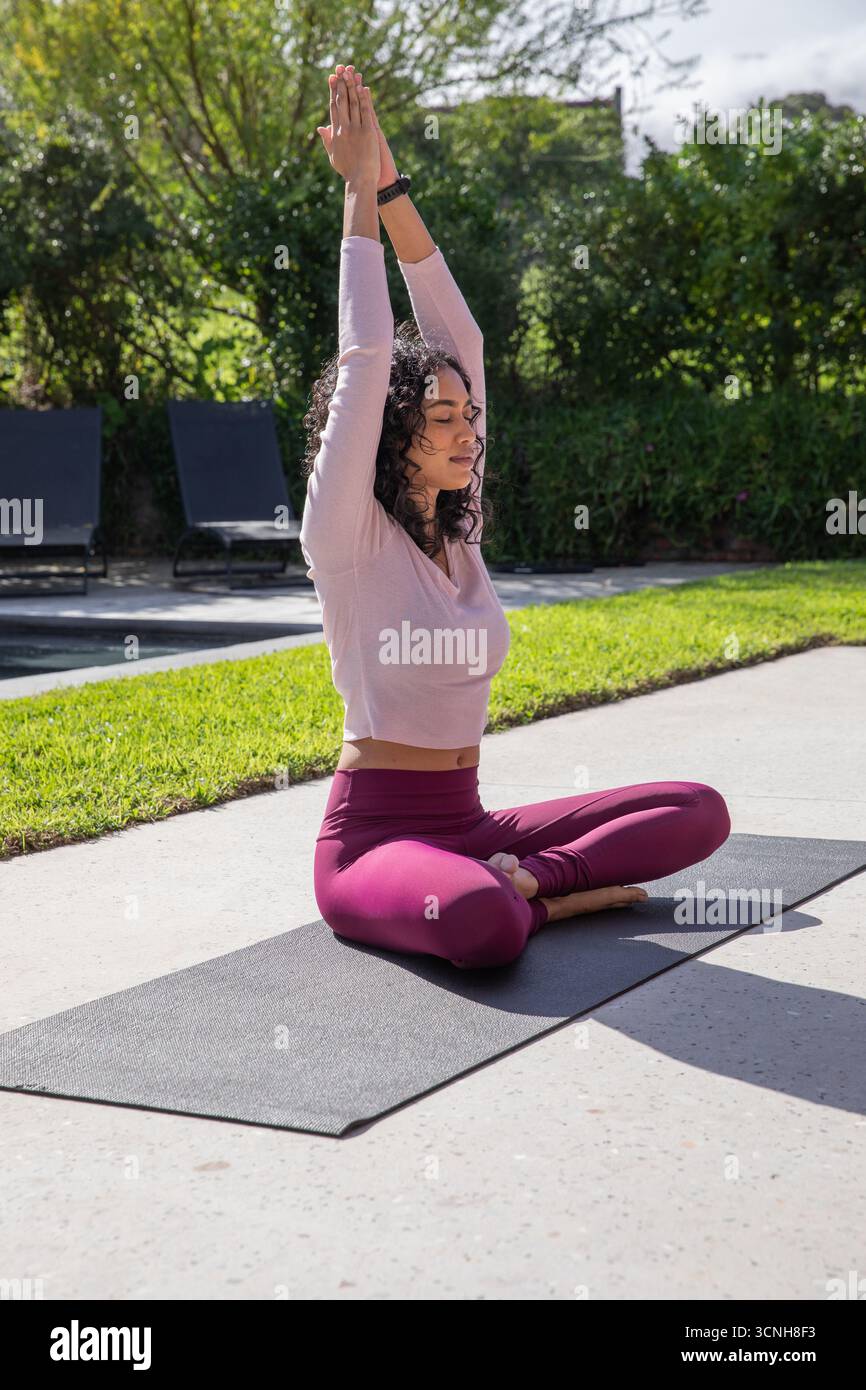 Donna che indossa un abbigliamento sportivo che pratica yoga sul patio vicino a poltrone con smartwatch e tappetino per yoga Foto Stock